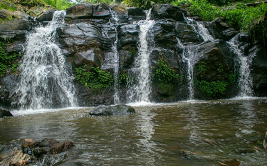 A scenic tropical waterfall cascading over dark volcanic rocks, surrounded by lush green bamboo and dense rainforest vegetation in Indonesia. Captured with a long exposure technique, the silky flowing © Eriec