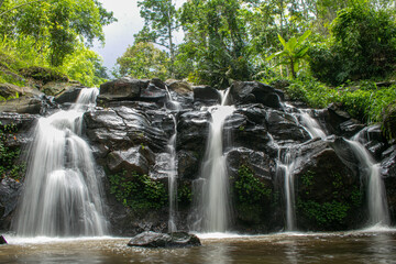 A scenic tropical waterfall cascading over dark volcanic rocks, surrounded by lush green bamboo and dense rainforest vegetation in Indonesia. Captured with a long exposure technique, the silky flowing © Eriec