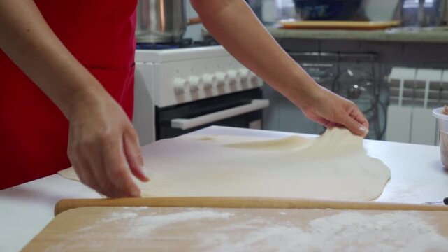 Close view of hands gently stretching thin dough over floured wooden countertop in home kitchen beside window, with soft natural light, visible flour dusting, smooth texture, and calm homemade