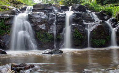 A scenic tropical waterfall cascading over dark volcanic rocks, surrounded by lush green bamboo and dense rainforest vegetation in Indonesia. Captured with a long exposure technique, the silky flowing © Eriec