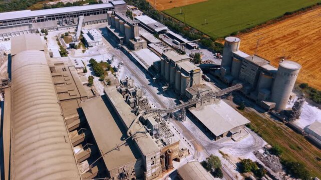 Large industrial cement plant with multiple storage silos. The high angle drone perspective shows the factory complex adjacent to agricultural fields.