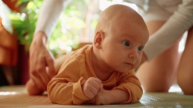 Young baby boy doing tummy time on a floor mat, developing neck muscles and learning to lift his head with gentle assistance from his caring mother in a sunlit room at home
