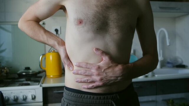 Bare chested man at home in the kitchen suffering from severe abdominal pain, holding his side and stomach with his hands due to a painful spasm, cramp, or digestive problem. Attack of cholecystitis
