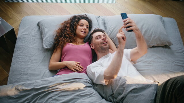 Couple lying bed browsing phone laughing together. Fun weekend morning at home.