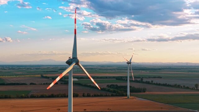 Wind turbines over farmland at golden hour. Warm side light outlines red marked blades above fields, distant hills, and a layered cloudy sky.