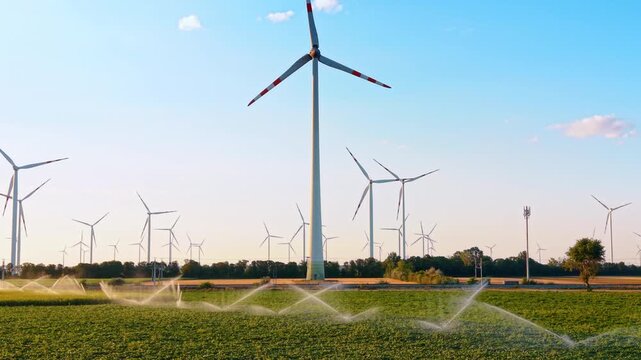 Wind turbine above irrigated green crops. Foreground sprinklers spray across a field beneath red marked blades and many distant towers.
