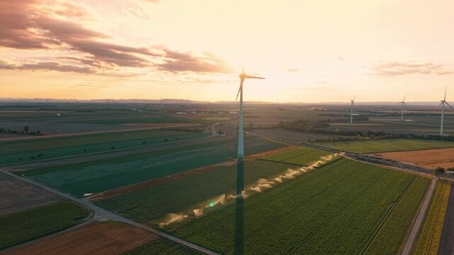 Wind farm and irrigated fields at sunset. Low sunlight casts a long turbine shadow across green crops beside spraying irrigation lines.