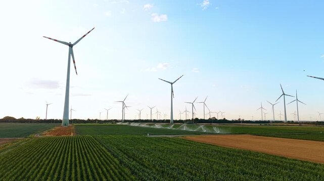 Wind turbines and crop rows across farmland. Parallel green plantings lead toward irrigation spray and numerous towers beneath clear evening light.
