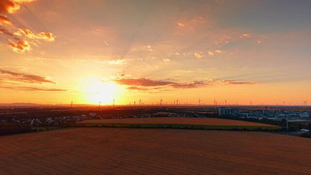 Aerial view of sunbeams at sunset over fields and wind farm. Sun rays break through orange clouds above a vast rural landscape with distant industrial structures.