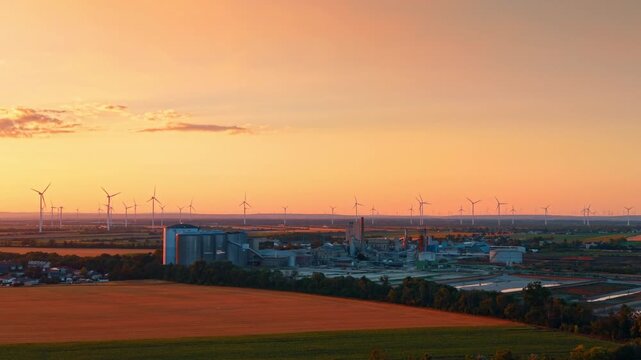 Aerial view of factory and wind farm across agricultural fields. Industrial facility sits surrounded by cultivated land under a warm gradient twilight sky.