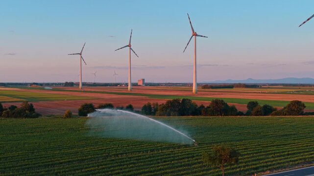 Curved water spray from irrigation system near wind turbines. A sprinkler system throws a large arc of water over green crops under a twilight sky.
