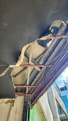 A cornice structure with its underside painted dark grey or black, but largely broken and fragmented, revealing a rusty metal frame system, insulation materials, and dangling cables through the damage © cagatay