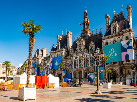Wide view of the H&ocirc;tel de Ville in Paris under a clear blue sky, its facade decorated with large "Paris 2024" Olympic and Paralympic banners, palm trees, event tents, and setup equipment in the square