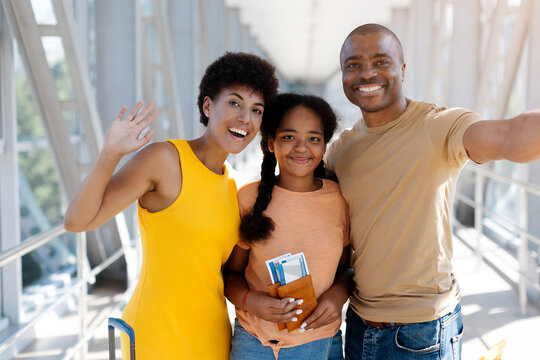 Cheerful Black family posing for smartphone selfie at the airport, mother waving and daughter displaying passports and boarding passes before their holiday flight