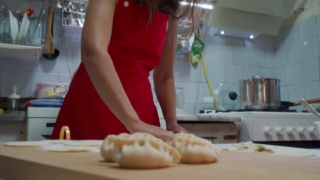 Hands of home cook shaping raw dumplings on wooden countertop with flour dusting, red apron visible on torso, tiled backsplash, stove and pot in background, warm indoor light and focused food