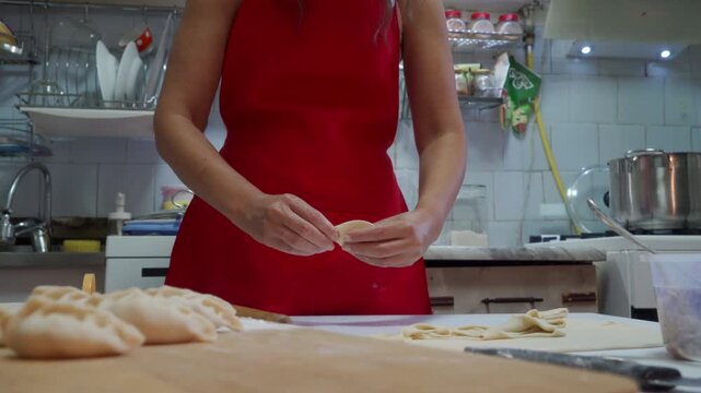 Midsection of woman in sleeveless red dress shaping dough into pastries on wooden countertop in home kitchen, with stainless pots, utensils, tiled backsplash, flour, and warm overhead light visible