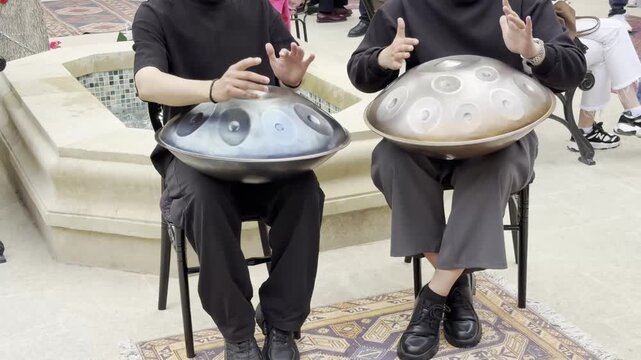 Two street musicians play hanga (handpan) during a city event. Performers sit by a small fountain and create atmospheric live music. A close-up of musical instruments and the playing process, the conc