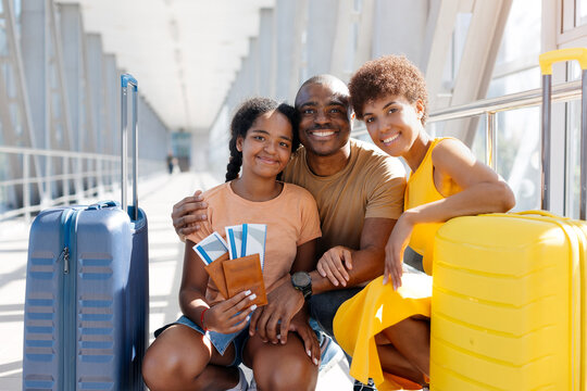 Smiling Black family of three posing closely between colorful suitcases in a bright airport terminal, with their daughter holding passports and boarding passes before departure