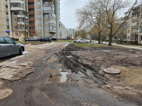 A muddy, damaged road with deep ruts and potholes filled with water in a residential area featuring Soviet-era apartment buildings. The foreground shows broken asphalt, mud tracks, and cracked