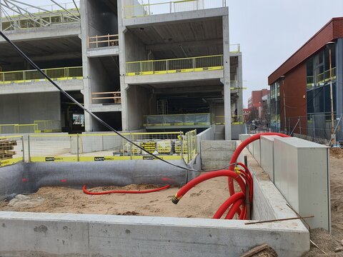 A construction site featuring a multi-story reinforced concrete building under active development. The structure displays exposed concrete columns, floor slabs, and open shafts with yellow safety