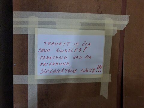 A close-up photograph of a handwritten threatening note in Lithuanian language taped to a brown wooden door or wall with masking tape. The message, written in red ink on white paper, aggressively
