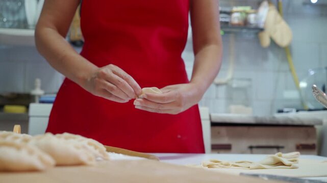 Close-up of hands forming dumplings from dough on wooden counter inside home kitchen, red apron visible from torso, flour dust on surface, tray of finished pieces nearby in warm natural light