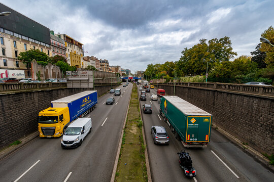 Aerial view from a bridge over a sunken multi-lane highway in Paris's suburbs, with a yellow "Help Transports" truck and a green "Gamba & Rota" truck among car traffic, cloudy sky, stone retaining wal