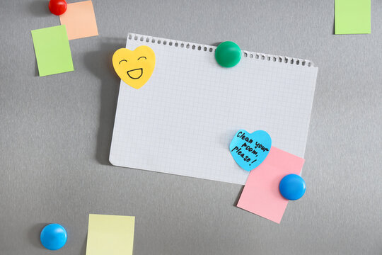 Blank grid paper notebook page with colorful sticky notes and magnets on a brushed metal refrigerator door, Various sticky notes in different colors and shapes, including a yellow heart with a smiley