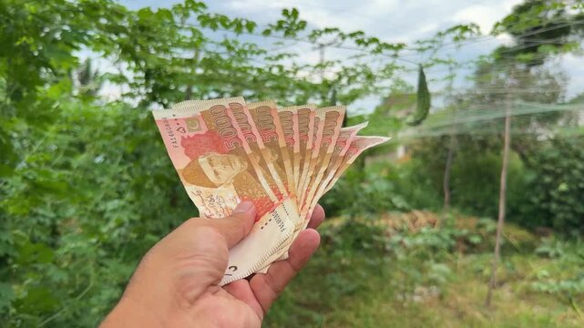 A farmer proudly holds up a 5,000 denomination Pakistani banknote against a wide, green crop field during harvest, symbolizing agricultural success, financial reward, and the dignity of rural labor.