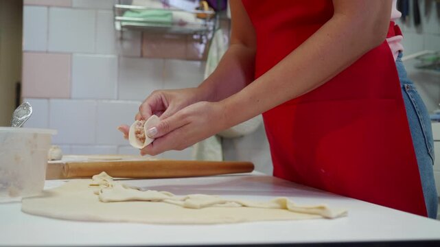 Home cook wearing red apron shaping fresh dough on white kitchen countertop, hands folding pastry beside rolling pin, measuring cup, flour, and pastry strips on work surface under warm indoor light