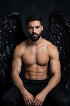 Shirtless muscular man with black feather wings poses against dark background. Athletic bearded guy strong fit torso, defined abs. Handsome male model looks like dark fallen angel mythical creature