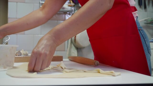 Close view of woman in red apron rolling thin dough on white countertop with wooden rolling pin, shaping pastry pieces in bright kitchen with tiled backsplash and clean home cooking atmosphere
