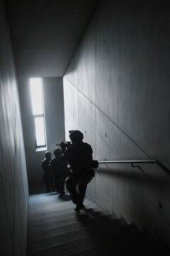 Group of soldiers in tactical gear climbing staircase inside concrete building. Military unit moving through hallway for mission.