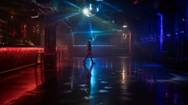 Man walking alone through empty nightclub under disco ball. Dark dance floor with blue and red neon lighting. Nightlife, clubbing and solitude concept in professional music venue.