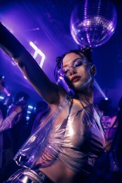 Young woman dancing at nightclub under disco ball. Stylish girl wearing trendy outfit and clear glasses enjoying electronic music party.