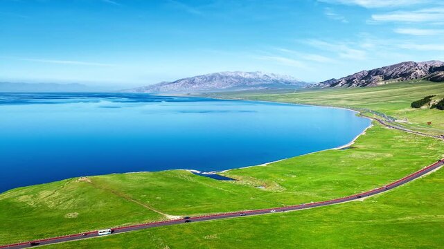 Aerial view of Sayram Lake with bright blue water, green grassland, scenic highway, and mountain background in Xinjiang, China.