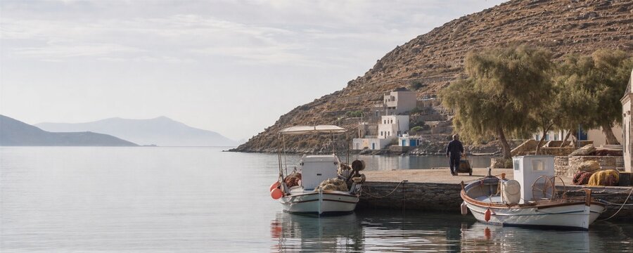 Fisherman pulling a cart near small traditional boats at a pier on a Greek island. Harbor scenery with calm blue water, white houses on a hill, and Mediterranean summer travel conc