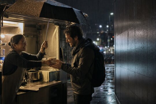 Woman street vendor selling hot soup to a man under an umbrella during a rainy night. Urban food stall service in city. Small business and meal delivery concept. Evening scene.
