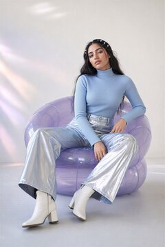 Stylish young woman sitting in modern inflatable armchair against white wall. Fashionable girl wearing blue sweater and silver metallic pants. Trendy home interior design studio co