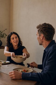 Woman serving food to man with hearing aid at dining table. Friends sharing meal during lunch at home. Inclusive communication, disability awareness and social connection concept.