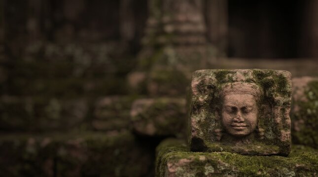 Close up of an ancient Khmer stone face carving covered in green moss among temple ruins