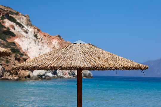A straw beach umbrella stands before turquoise water and rocky cliffs on a sunny summer day on Firiplaka beach in Milos, Greece.