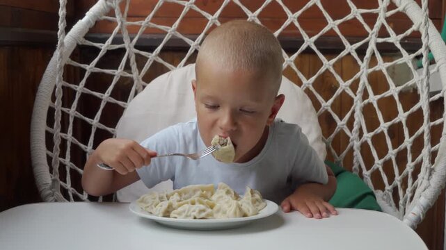 Blond toddler sitting in round woven hanging chair over white dining table, leaning toward large bowl of creamy pasta and using fork, warm indoor light, casual cozy home kitchen scene