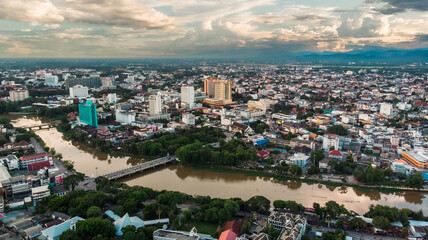Obraz premium Aerial drone view of Chiang Mai cityscape and Ping River with surrounding mountain range under a cloudy golden hour sky in Northern Thailand