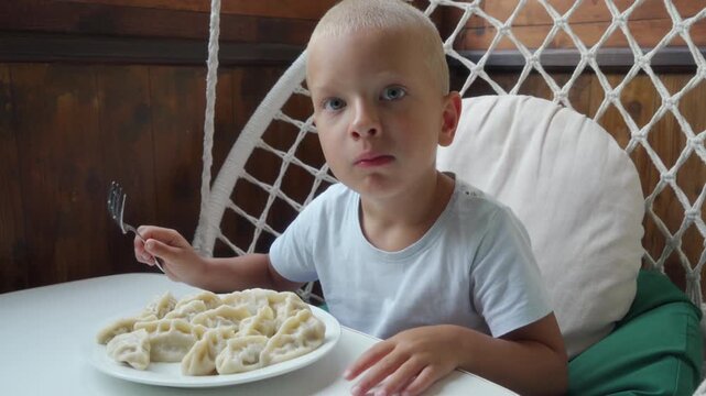 Child with shaved head sits in white wicker hanging chair, eating forkfuls of pasta from plate at home table, with neutral expression, soft light, wooden paneled background, relaxed domestic scene