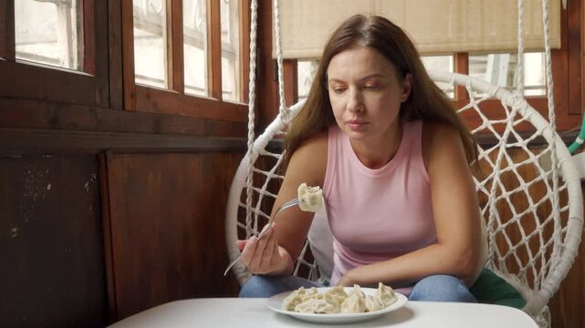 Young woman in sleeveless pink top sits in woven hanging chair beside small round table, eating salad from white bowl near wooden window with soft natural daylight and quiet urban cafe mood