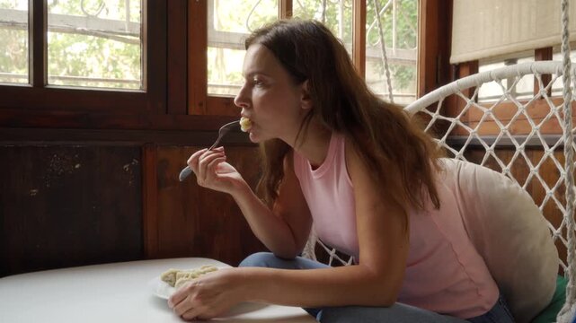 Young woman in casual sleeveless top leans forward on cushioned wicker chair to eat pastry beside large sunlit window, warm natural light illuminating face, wood frame, calm home setting