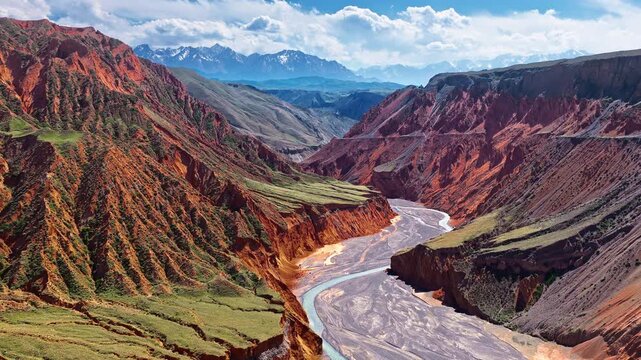 Aerial high-angle view of a dramatic canyon carved through colorful red rock formations with a winding river channel under a partly cloudy sky in Xinjiang, China.