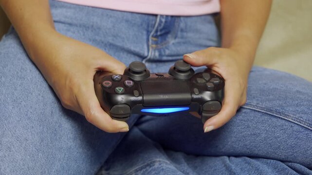 Closeup shows adult hands gripping wireless game controller while seated crosslegged on couch, wearing blue denim jeans and light top, warm indoor lighting highlighting textured plastic, glowing