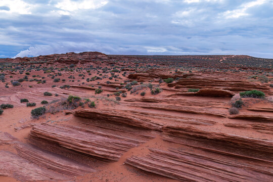 Sculptured red sandstone desert landscape at Horseshoe Bend near Page, Arizona. Layered rock formations and sparse vegetation. Mountains and cloudy sky in the background. 
